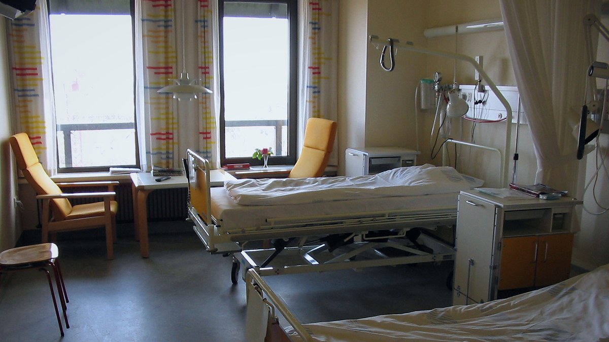 Habitación de hospital con dos camas articuladas, sillones y ventanas con cortinas