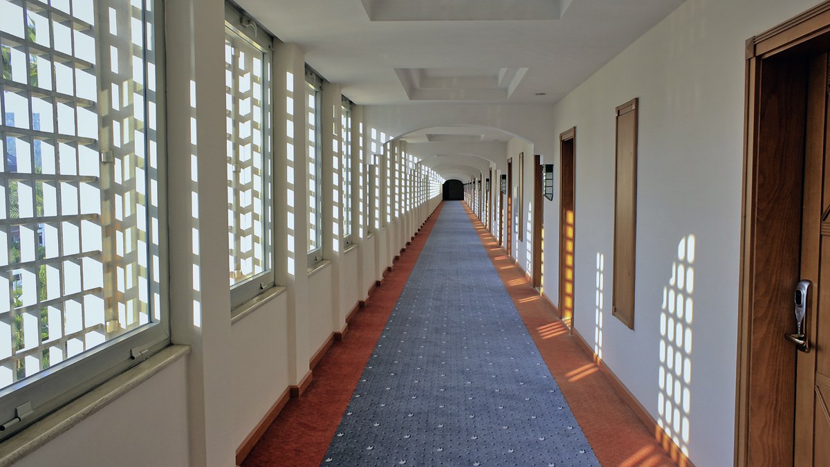 Pasillo largo y vacío de un hotel con moqueta, ventanales a un lado y puertas de habitaciones al otro