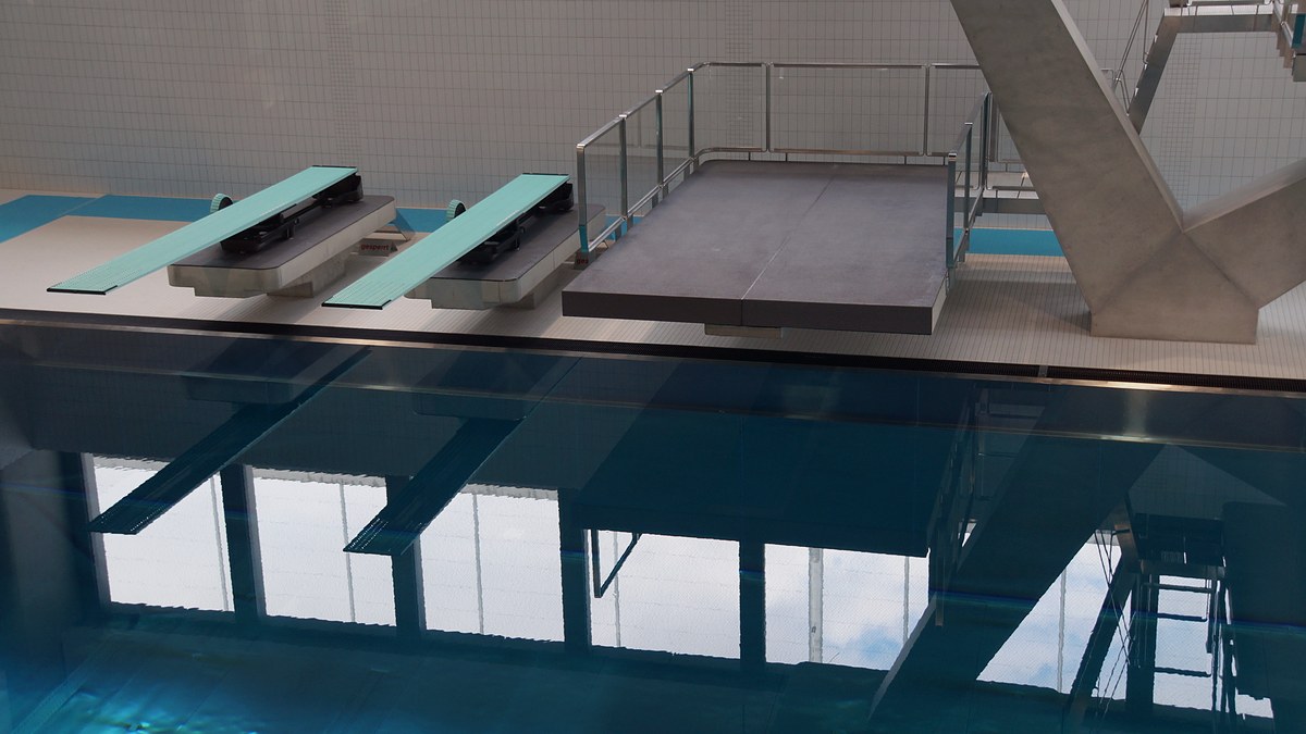 Piscina cubierta sin bañistas, con dos trampolines y una plataforma de saltos que se reflejan en el agua